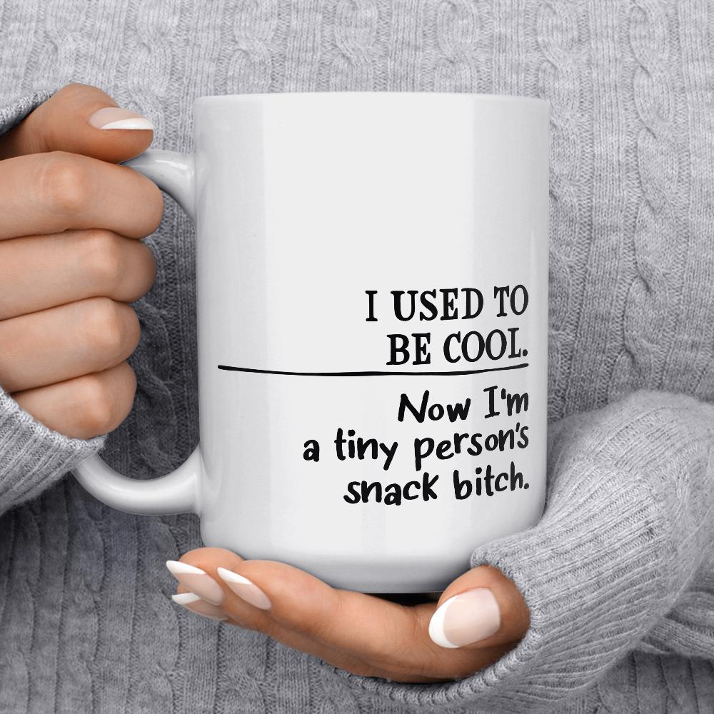 white mug that says I used to be cool, now I'm a tiny person's snack bitch being held by person wearing grey sweather