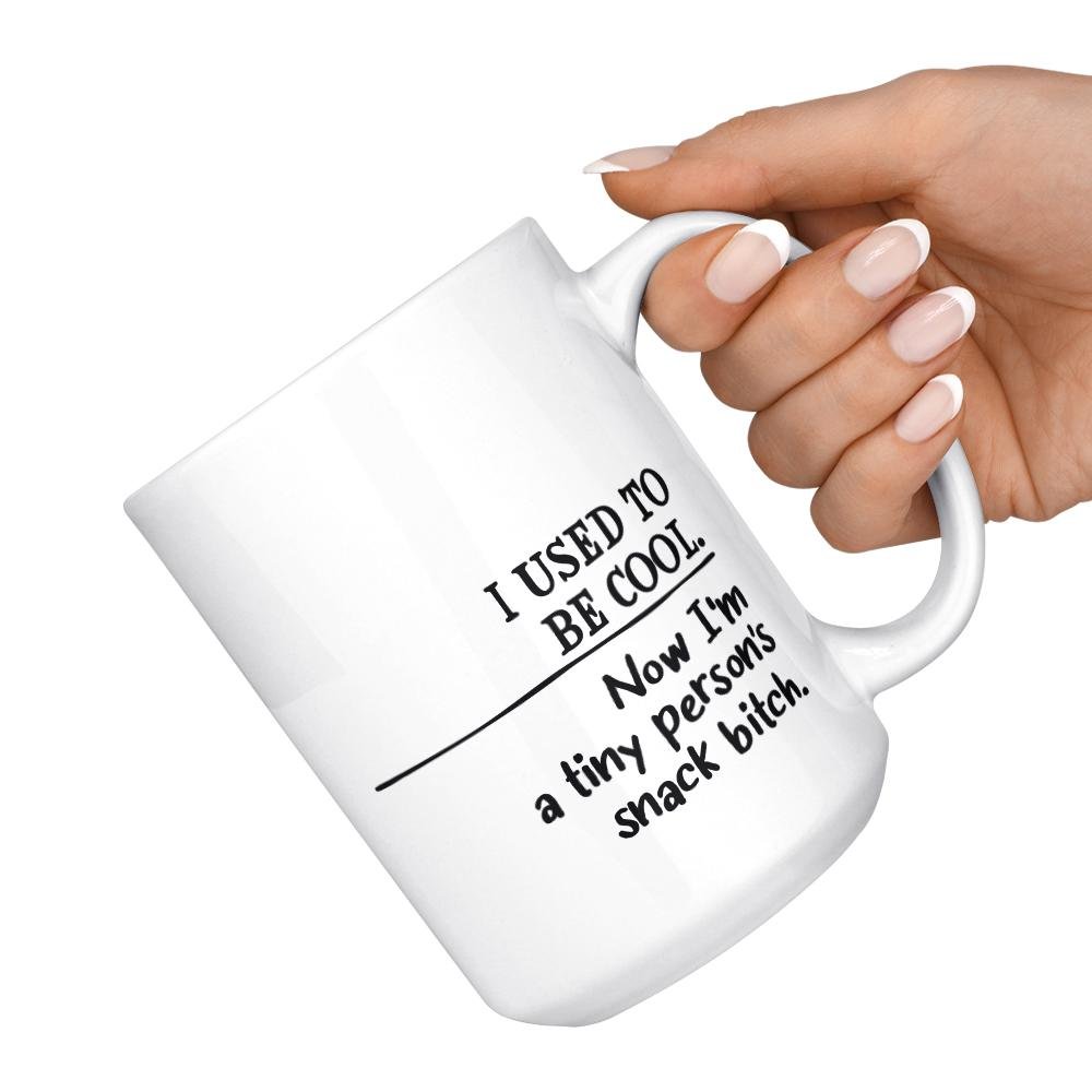 white mug that says I used to be cool, now I'm a tiny person's snack bitch being held by hand