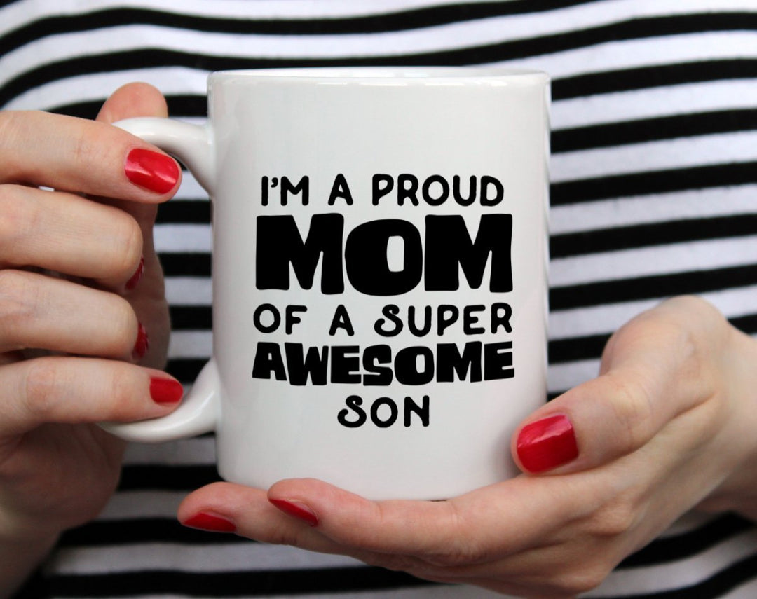 White mug that says I'm a proud mom of a super awesome son being held by woman wearing black and white top with red nail polish